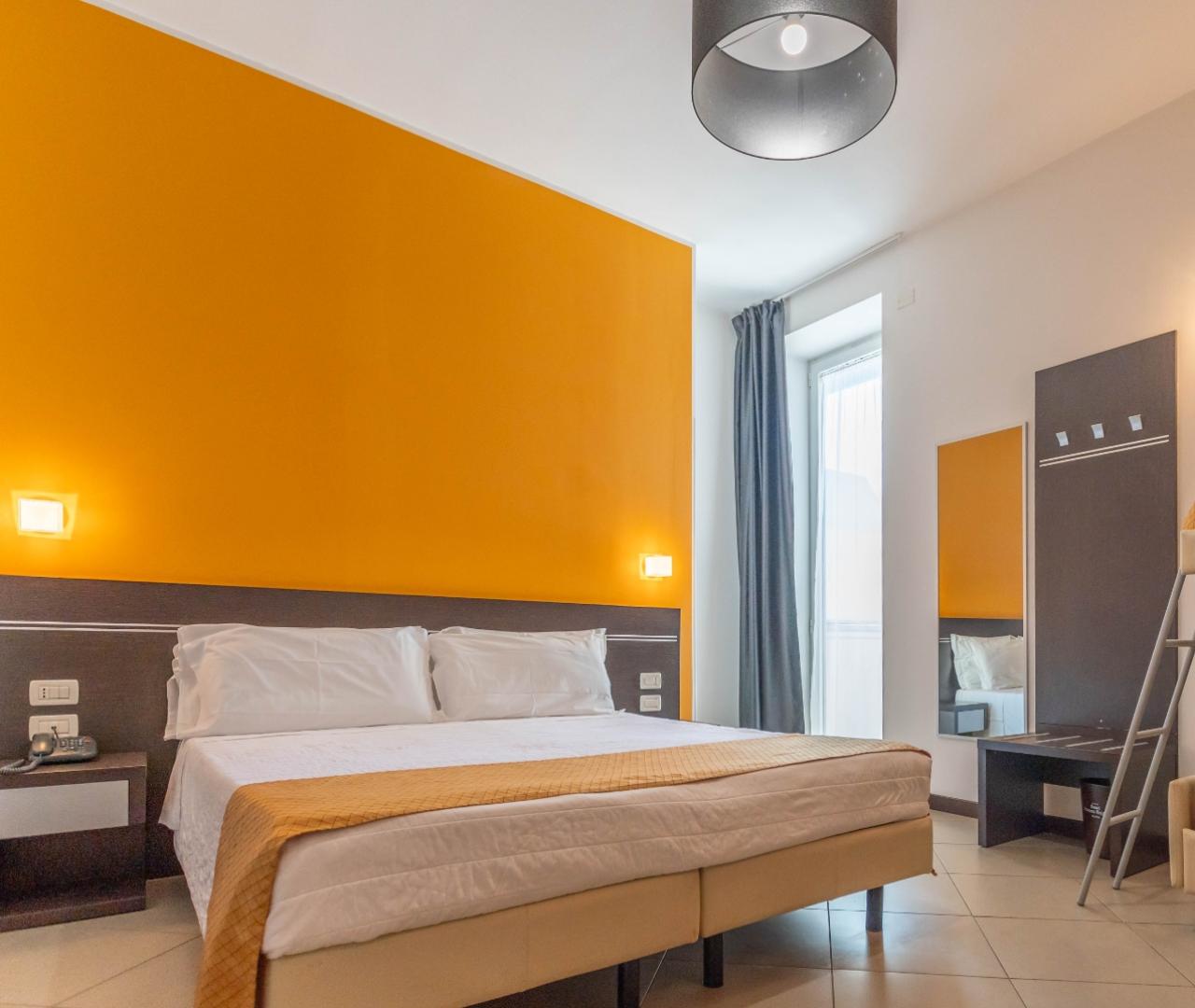Camera accogliente con parete arancione, letto matrimoniale e luce soffusa, atmosfera calda e rilassante.
