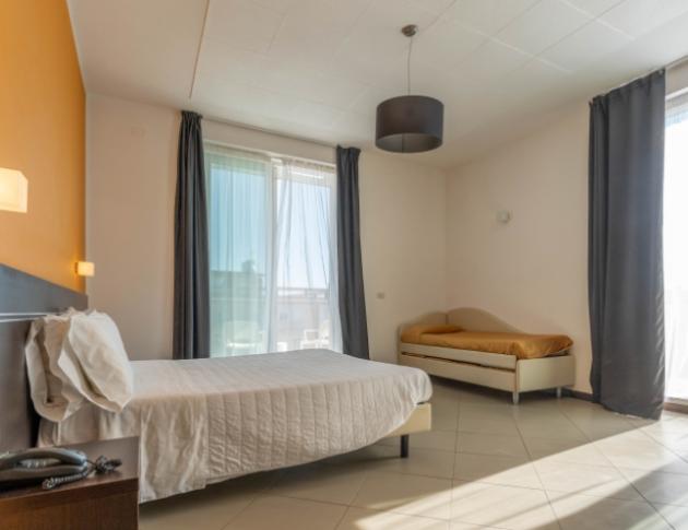 Camera luminosa, letto bianco, parete arancione, tende grigie, atmosfera accogliente e rilassante.